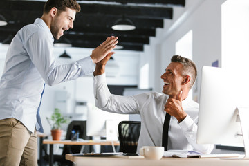 Fototapete - Photo of cheerful businessmen 30s in formal clothes giving high five together in office, during successful deal