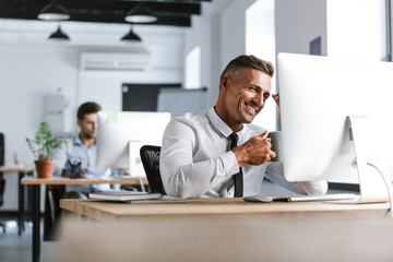 Fototapete - Photo of cheerful adult man 30s wearing white shirt and tie drinking tea from cup, while sitting by computer in office