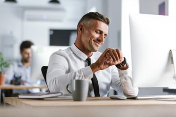 Fototapete - Image of adult businessman 30s wearing white shirt and tie sitting at desk in office by computer, and looking at wrist watch