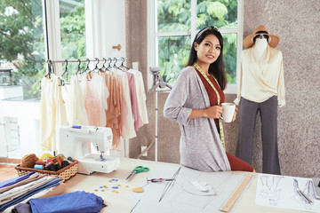 Charming Asian female smiling and looking at camera while holding mug of hot beverage and leaning on table in workshop