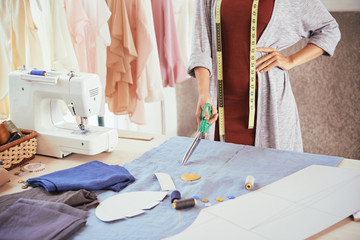 Anonymous fashion designer holding sharp scissors and standing near table with blue cloth while working in tailoring workshop