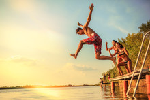 Group Of Friends Jumping Into The Lake From Wooden Pier.Having Fun On Summer Day.