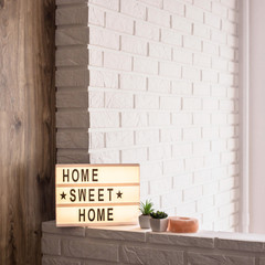 Home sweet home - it is written on a decorative frame with illumination in the interior between a white brick wall and a brown wooden one next to succulents and a pink candlestick. Copyspace