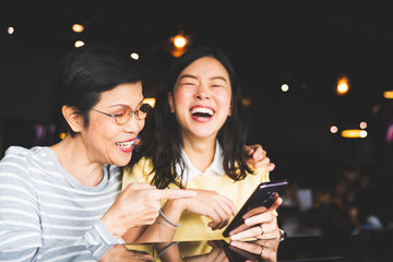 Asian mother and daughter laughing and smiling on a selfie or photo album, using smartphone together at restaurant or cafe, with copy space. Family love, holiday activity, or modern lifestyle concept