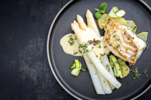 Modern German Fried Cod Fish Filet With White Asparagus In Hollandaise Sauce With Roast Potatoes And Sliced Zucchini As Top View On A Plate With Copy Space Left