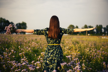 Wall Mural - View from the back of a woman brunette with a bouquet of purple flowers on a flower field in the sunset, freedom, dreams