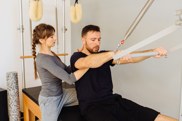 Young sporty female instructor of pilates helping beard man workout in cadillac bed. Two people working in pilates studio, woman assistant supporting and correcting male patient beginner.