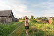 a model girl in a retro dress is holding in her hands a vintage suitcase on an abandoned country road overgrown with grass between the village courtyards and huts