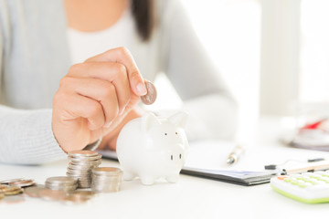 Wall Mural - Woman hand putting money coin into piggy bank with stack of coins. Computer calculator on office table. Saving money wealth and financial concept.