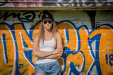 Fototapeta  - Portrait of young beautiful woman wearing white tank shirt and blue jeans and black hat on brick wall with graffiti background