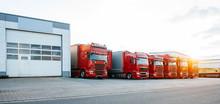 View Of Squadron Group Of New Red Cargo Trucks Parked In A Row Near Warehouse Building In Bright Sunset Light