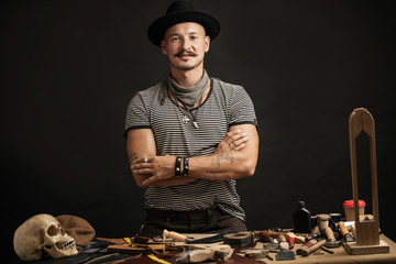 Wall Mural - Moustached craftsman with creative capon his head on, posing near his working place, at the table with crafting tools and rough leather in studio. Small Business, Cobblering, Leather Workcraft