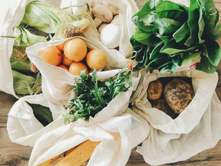 fresh vegetables in eco cotton bags on table in the kitchen. lettuce, corn, potatoes, apricots, bananas, rucola, mushrooms from market. zero waste shopping concept.   ban plastic