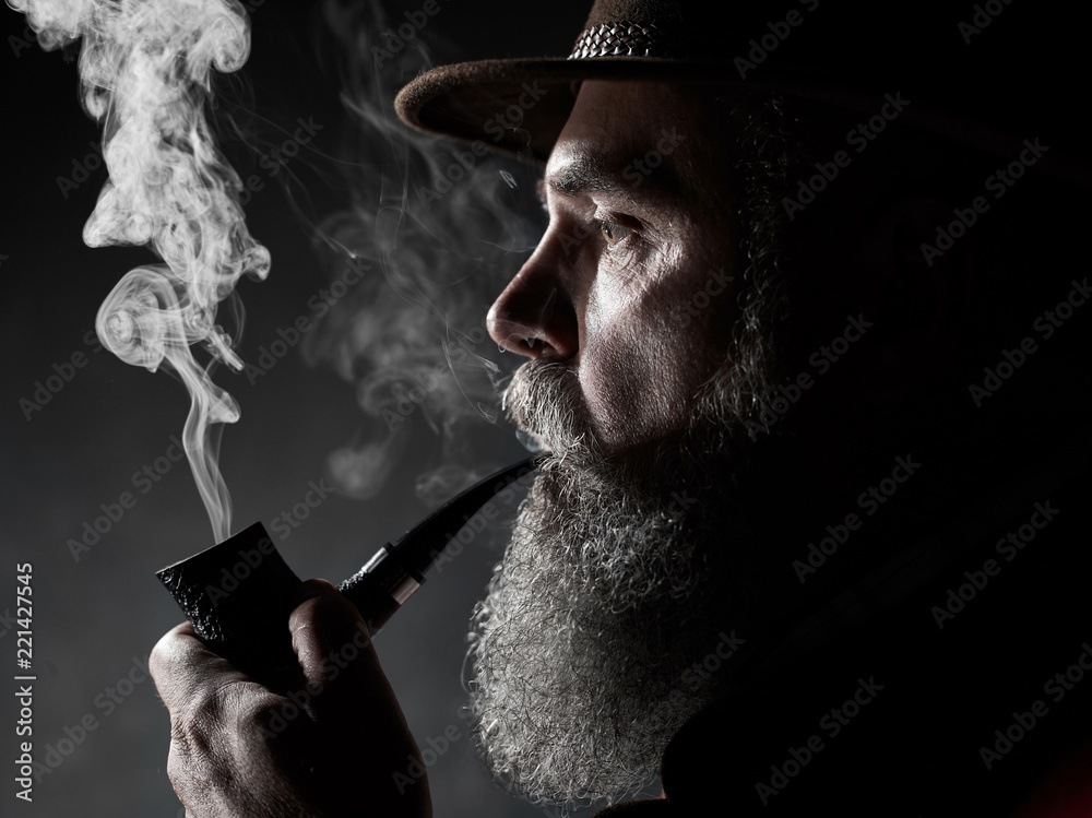 dramatic portrait of senior man in hat smoking tobacco pipe. Profile view of Austrian, Tyrolean, Bavarian old man - obrazy, fototapety, plakaty 