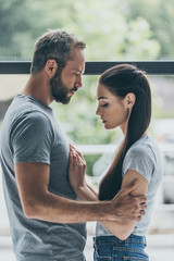 Wall Mural - side view of bearded man embracing sad young girlfriend while standing together near window