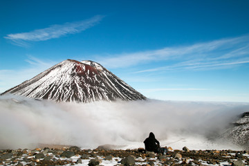 Tramper having rest in front of active volcano, mountain hiker and climber having snack and enjoys spectacular view of Mount Ngauruhoe peak above clouds, Tongariro National Park, New Zealand