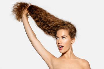 Young woman holding hair in hand on white background. Good-looking girl with healthy long wavy hairstyle. Beauty and haircare concept.