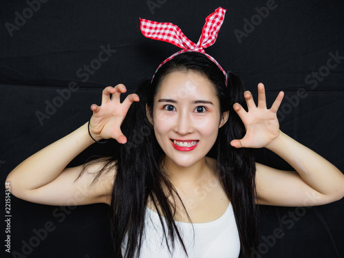 Fotografía Portrait of sexy Chinese woman with long black hair, pink hair ribbon and white halter against black background, smiling at camera and make threatening gestures