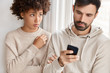 © WHstudio Leushin N - Jealous black girlfriend tries to peep at boyfriends smart phone, feels sad as he texts with someone, dressed in casual sweaters, have troubles with relationships. Multiethnic couple indoor at home
