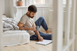 © WHstudio Leushin N - Relaxed bearded Caucasian guy enjoys spare time, calm atmosphere at home, holds mug with coffee, reads interesting novel, looks attentively at book, sits on floor near not made bed. Hobby concept