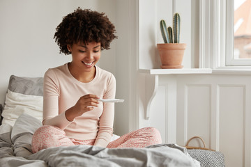 Happy future mother looks at control line of pregnancy test, dressed in nightwear, rejoices good news, sits crossed legs on comfortable bed. Pregnant beautiful woman at home. Maternity concept