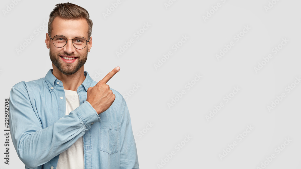 This idea just fascinated me. Attractive young unshaven individual with happy expression, indicates with index finger at blank copy space, advertises you place to vist, isolated over white wall - obrazy, fototapety, plakaty 