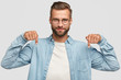 Lets go downstairs. Photo of attractive unshaven man with appealing look, points down with both index fingers, dressed in casual shirt, isolated over white background. Advertisement concept.