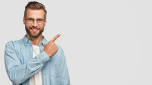 This Idea Just Fascinated Me. Attractive Young Unshaven Individual With Happy Expression, Indicates With Index Finger At Blank Copy Space, Advertises You Place To Vist, Isolated Over White Wall