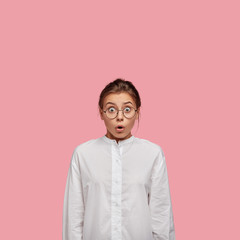 Wall Mural - Vertical shot of terrified young woman with shocked expression, stares with unexpected gaze, opens mouth from wonder, wears casual white shirt, isolated over pink background. Girl in stupor.