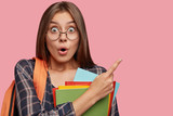 Fototapeta  - Horizontal shot of stupefied brunette pupil has surprised scared expression, points at upper right corner with index finger, dressed in checkered shirt, carries dictionary, going in school library.