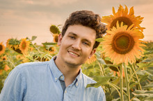 A Close Up Portrait Of A Young Smiling Man In A Field Of Sunflowers. Against The Backdground Of The Setting Sun And Orange Flowers.