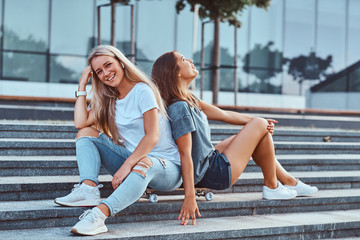 Wall Mural - Portrait of a two young hipster girls sitting together on skateboard at steps on a background of the skyscraper.