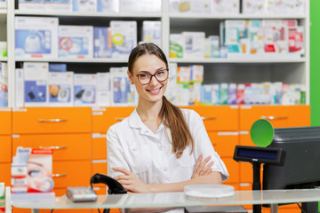 Wall Mural - A youthful pleasant dark-haired girl with glasses,dressed in a medical overall,greets visitors at the cash desk in a new pharmacy.