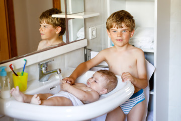 Cute adorable baby taking bath in washing sink and grab water tap. Kid boy helping and playing with sister. Happy brother and child having fun. Healthy children in bathroom together. Family time.