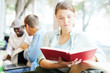 © pressmaster - Young woman sitting by window with open book and reading it with her groupmates preparing for project on background