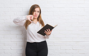 Wall Mural - Young adult woman standing over white brick wall reading a book with angry face, negative sign showing dislike with thumbs down, rejection concept