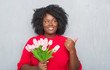 © Krakenimages.com - Young african american woman over grey grunge wall holding pink flowers bouquet pointing and showing with thumb up to the side with happy face smiling