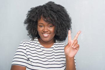 Canvas Print - Young african american plus size woman over grey grunge wall smiling with happy face winking at the camera doing victory sign. Number two.