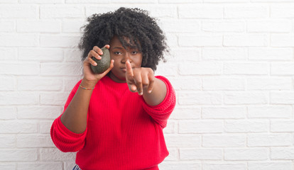 Wall Mural - Young african american woman over white brick wall eating avocado pointing with finger to the camera and to you, hand sign, positive and confident gesture from the front