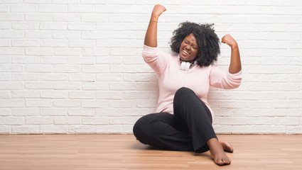 Poster - Young african american woman sitting on the floor wearing headphones celebrating surprised and amazed for success with arms raised and open eyes. Winner concept.