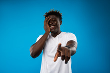 Are You Kidding Me. Waist Up Portrait Of Young Man Pointing At You And Laughing. He Is Wearing White T-shirt And Touching Face. Isolated On Blue Background