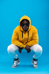 Street fashion. Studio portrait of young man in stylish yellow hoodie sitting on haunches and looking at camera with calm expression