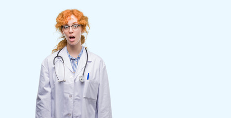 Canvas Print - Young redhead woman wearing doctor uniform afraid and shocked with surprise expression, fear and excited face.