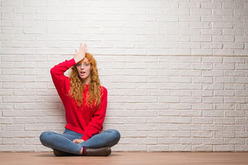 Wall Mural - Young redhead woman sitting over brick wall surprised with hand on head for mistake, remember error. Forgot, bad memory concept.