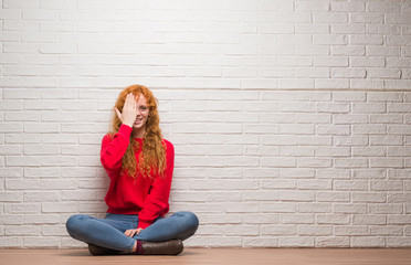 Canvas Print - Young redhead woman sitting over brick wall covering one eye with hand with confident smile on face and surprise emotion.