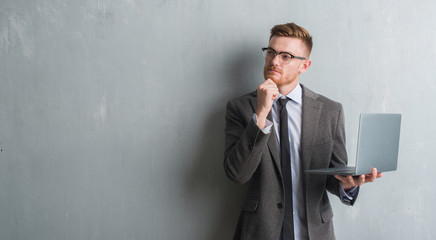 Wall Mural - Young redhead elegant business man over grey grunge wall using laptop serious face thinking about question, very confused idea