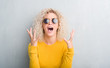 Young blonde woman with curly hair over grunge grey background crazy and mad shouting and yelling with aggressive expression and arms raised. Frustration concept.