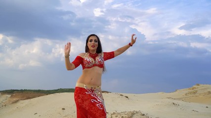 Wall Mural - Oriental Beauty dancing sensual belly dance outdoors. Attractive girl in a red dress is dancing in the desert in the background sky with clouds.