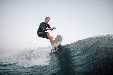 Wall Mural - Active man wakesurfing on the board down the blue wave against the background of clear sky