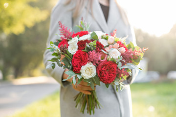 Lovely woman holding a beautiful autumn wedding bouquet. flower arrangement with white and red garden roses. Green lawn on background. Bright dawn or sunset sun